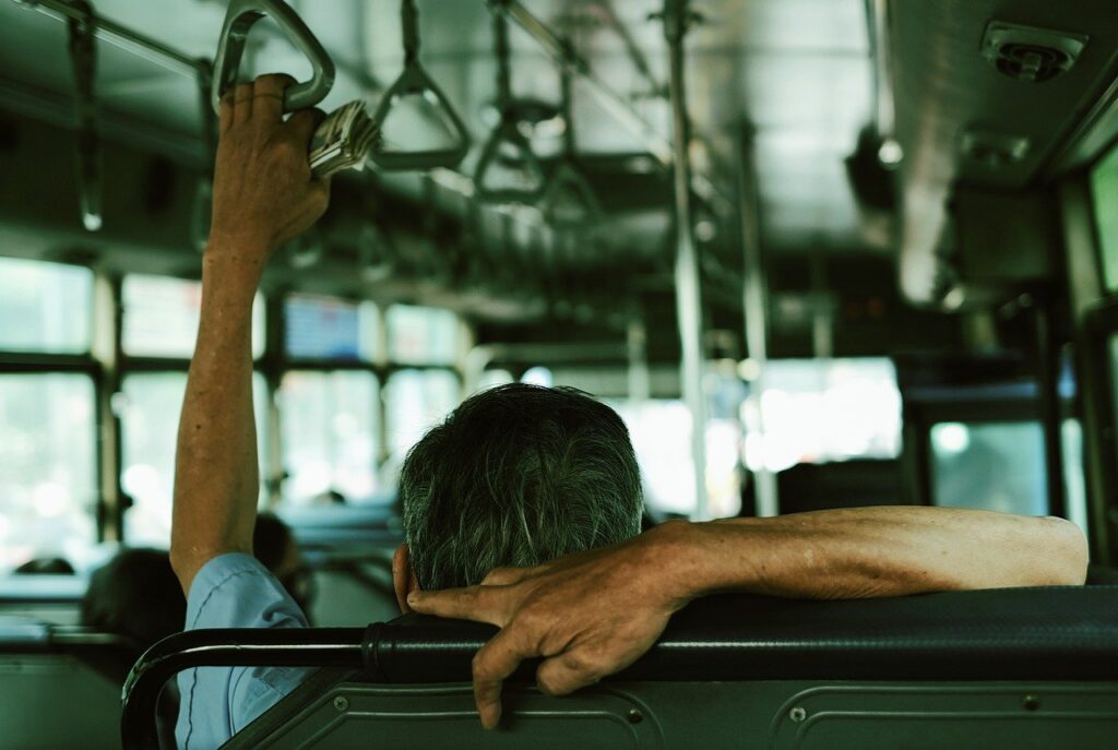 A man with gray hair sits on a bus while holding the support ring 