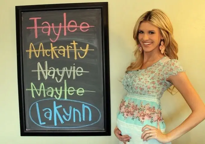 a chalkboard with the names “taylee” “mckarty” “nayvie” and “maylee” crossed out, with “lakynn” circled. a pregnant woman stands next to it