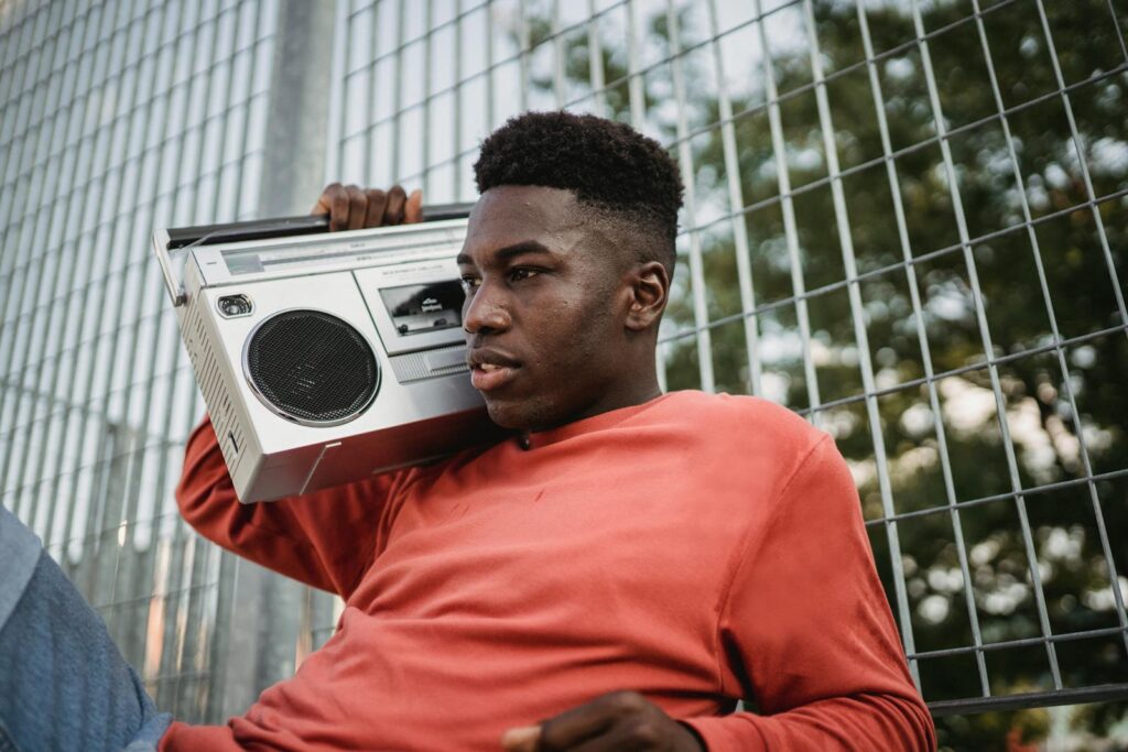 Young pondering African American male listening to radio from old tape recorder while looking forward near grid fence in town