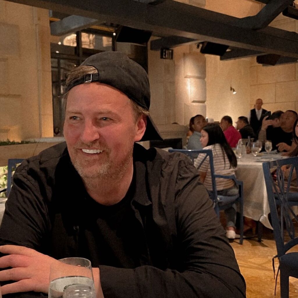 Matthew Perry smiling with a baseball cap on