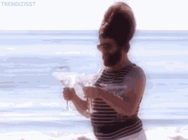 A gif of a man with a beard walking on the beach carrying champagne glasses 