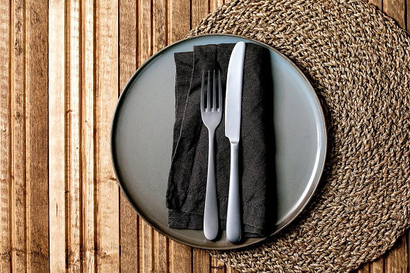 Empty ceramic plate with fork and knife on textile napkin on knitted straw round napkin over wooden plank background. Flat lay. space