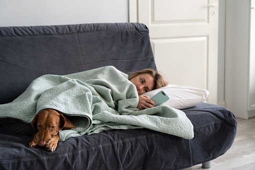 Sad woman lying on sofa with dog, using smart phone wait for boyfriend call or text, feels depressed