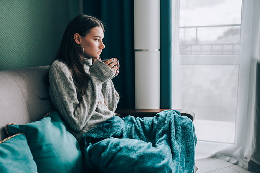 No central heating, cold apartment. Young brunette woman drink hot tea, look out the window, wrapped in a blanket in cold apartment