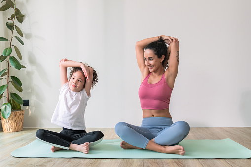 Healthy Latin American mother and her elementary-aged kid stretching their arms on an exercise mat during yoga class together at home for body care with wellbeing and positive emotion.