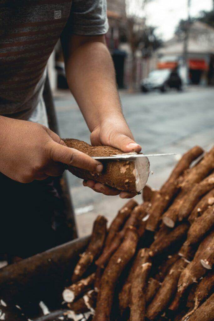 A person peeling cassava with a knife at an outdoor market, showcasing traditional food preparation.