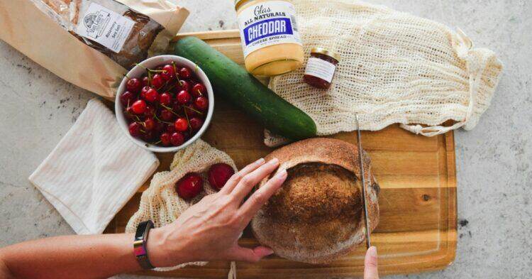 Person cutting bread on cutting board with groceries scattered on the table (Unsplash | Grace O'Driscoll)
