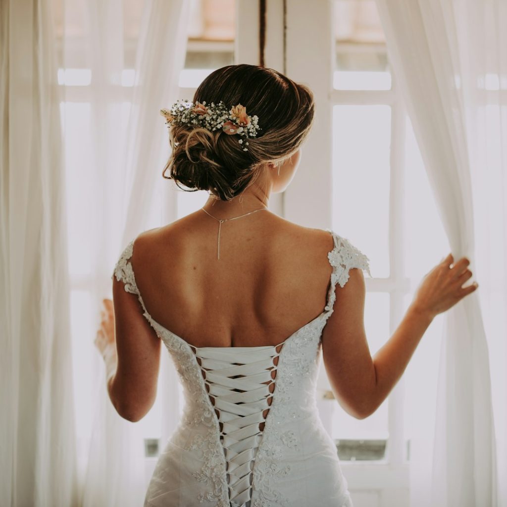 woman in wedding dress standing near window looking outside while holding the curtains