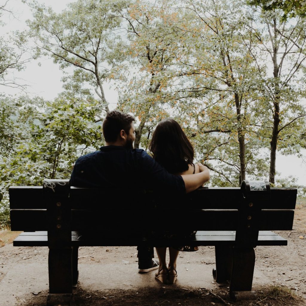 man looking to woman sitting on black wooden bench in front of tall trees during daytime
